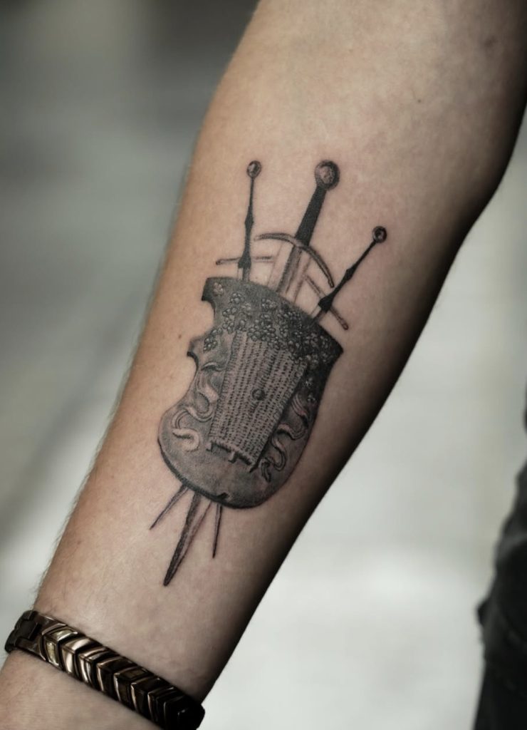 Tattoo van een gedetailleerd schild met zwaarden op de onderarm, door Mitchel.