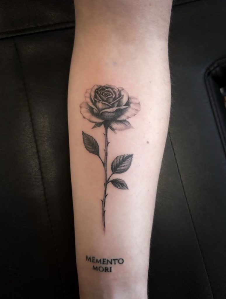 Zwart-grijze roos met steel en de tekst Memento Mori op de onderarm, door Mitchel.