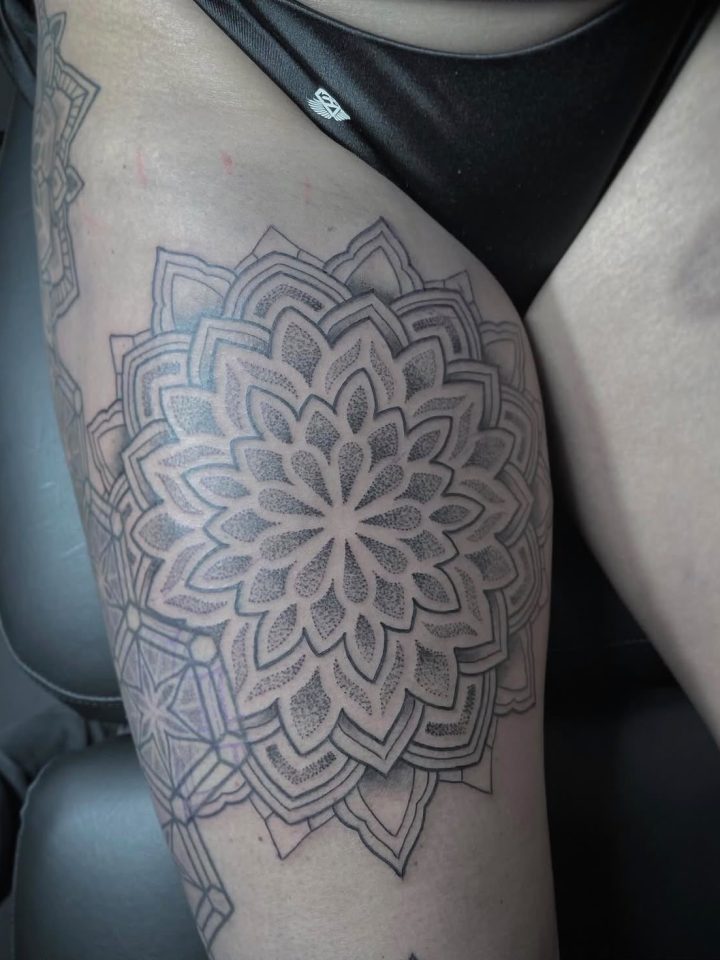 Mandala tattoo with layered floral shapes, fine lines and dots on a thigh, by Kenneth.