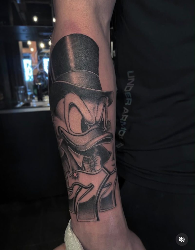 Comic tattoo van een eend met hoge hoed in zwart en grijs op een onderarm, door Kenneth.