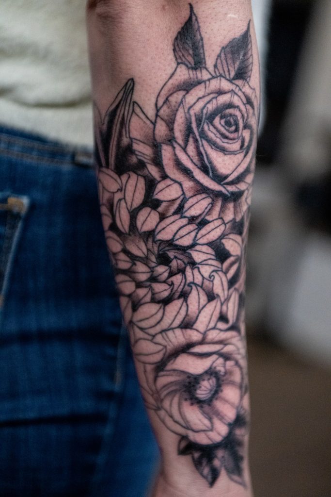 Lijnwerk tattoo van een roos met bloemen en bladeren op een onderarm, door Roger.