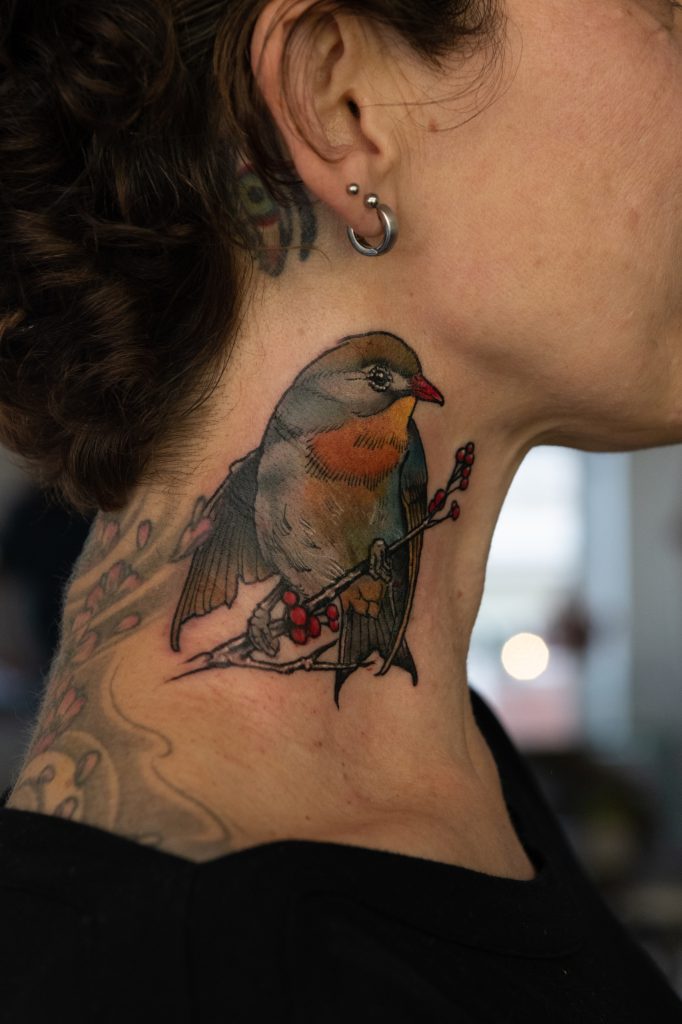 A colourful bird on a branch with red berries in Lucie’s neck tattoo.