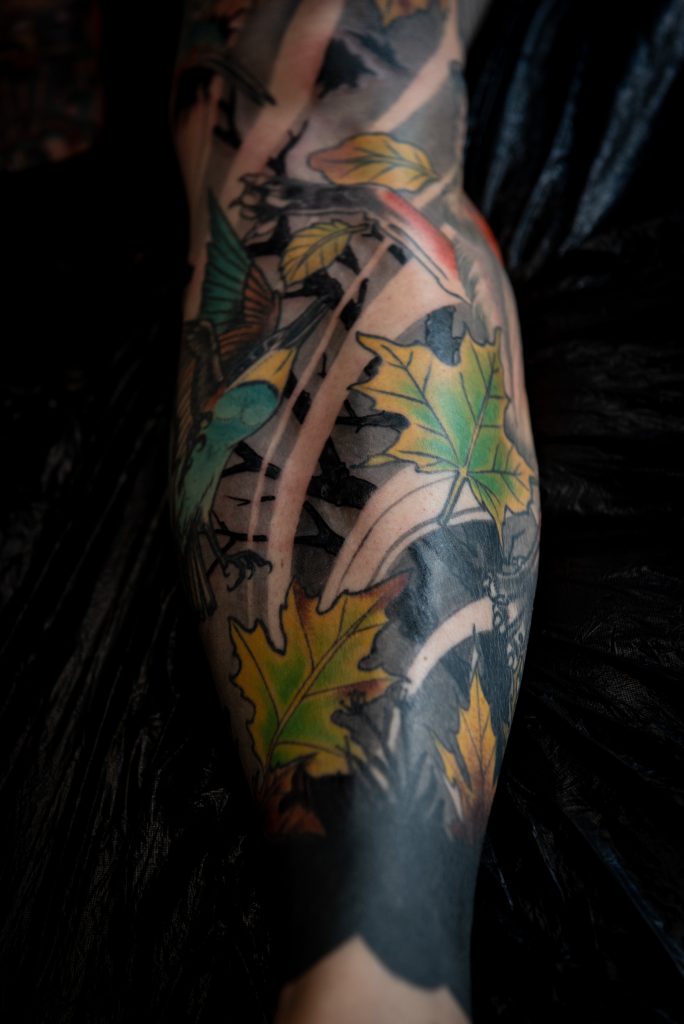 Detail of Lucie’s lower-leg tattoo with colourful birds, autumn leaves and a dark background.